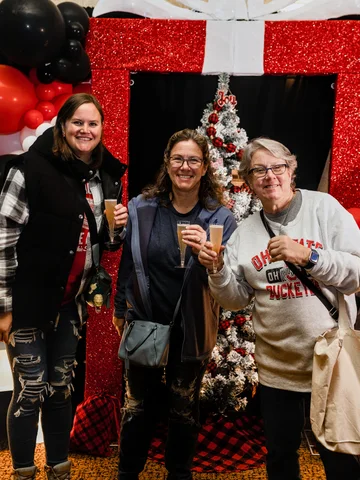 Three guests hold drinks in front of a red gift-shaped photo backdrop at the 2023 Ohio Made Holiday Market.