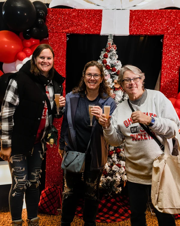 Three guests hold drinks in front of a red gift-shaped photo backdrop at the 2023 Ohio Made Holiday Market.