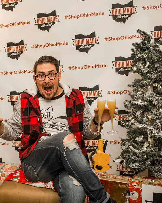 A volunteer in a plaid vest holds two glasses of orange drinks in front of the step and repeat wall.