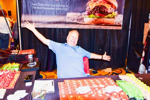 A vendor with outstretched arms smiles behind a table stacked with colorful boxes.