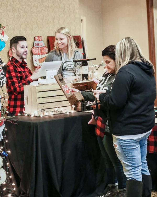 Two people in red plaid ring up a customer at a crate table with string lights at the 2019 Ohio Made Holiday Market.