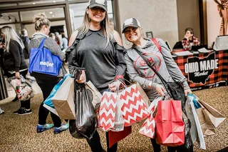 Two shoppers in ball caps smile with their arms full of colorful shopping bags.