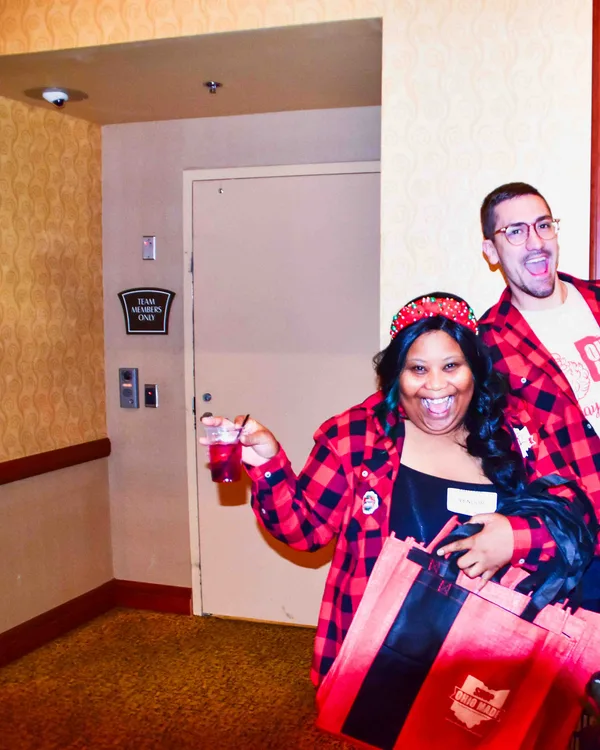 Two volunteers in plaid pose, one holding a drink and a large tote.