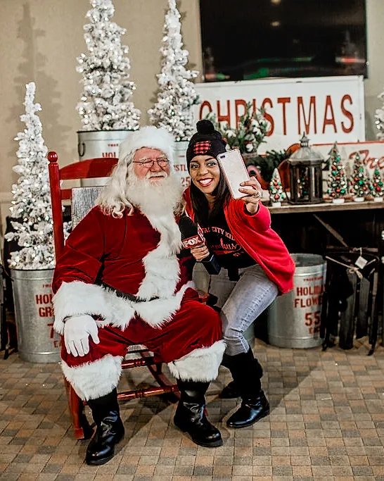 Santa sits in a rocking chair beside a woman taking a selfie, with Christmas tree signs behind them.