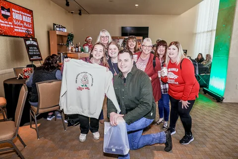 A group of guests poses in a lounge while one holds up a sweatshirt, with a drink specials sign behind them.
