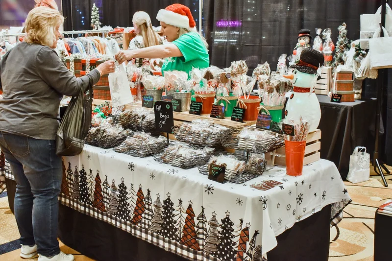 A maker in a Santa hat hands a bagged gift across a table of treats to a shopper at the 2023 Ohio Made Holiday Market.