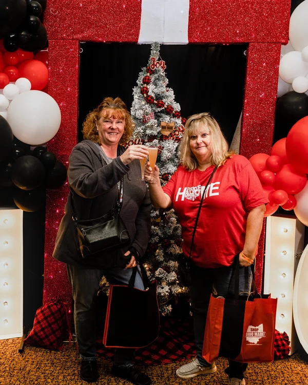 Two guests raise their drinks in a toast in front of the red gift photo set at the 2023 Ohio Made Holiday Market.