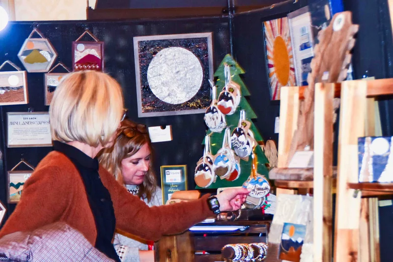 A shopper reaches for a handmade ornament on a table of mountain landscape art at the 2024 Ohio Made Holiday Market.
