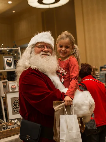 Santa in a red velvet suit holds a smiling young girl in a red top beside a table of framed owl art, a white shopping bag in front of them.
