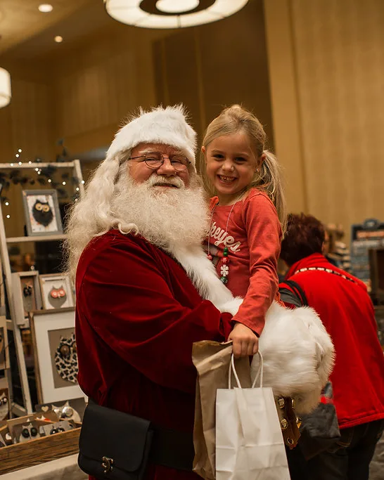 Santa in a red velvet suit holds a smiling young girl in a red top beside a table of framed owl art, a white shopping bag in front of them.