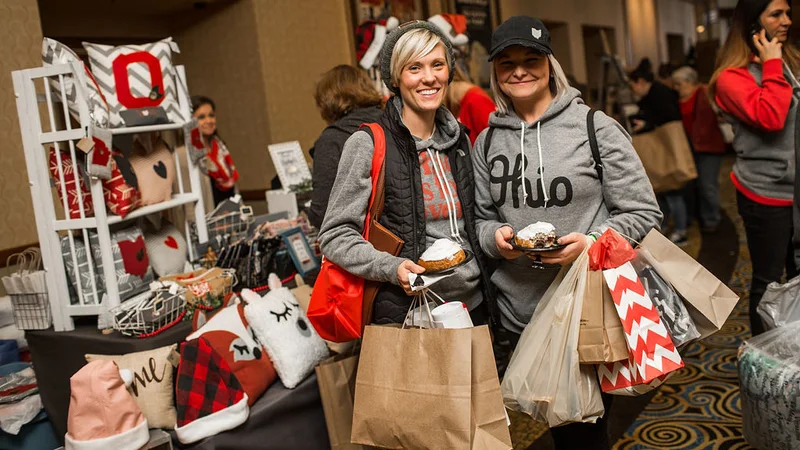 Two shoppers in Ohio sweatshirts carry shopping bags at a maker booth.
