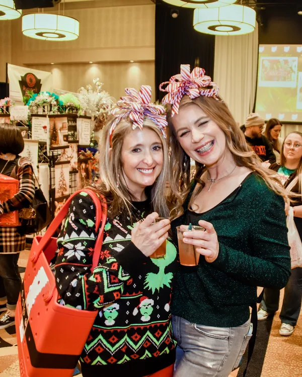 Two shoppers in festive sweaters wear bows on their heads and hold small drinks and a red tote.