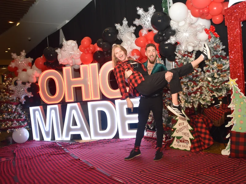 A volunteer in red plaid is lifted into the air by a friend in front of the glowing Ohio Made marquee letters at the 2025 Ohio Made Holiday Market.