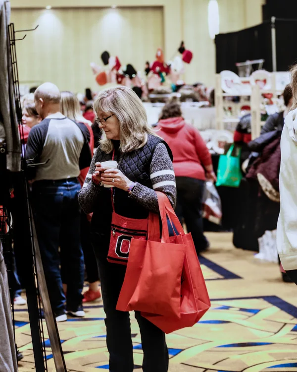 A shopper in a vest carries a red tote bag and a cup at the 2019 Ohio Made Holiday Market.